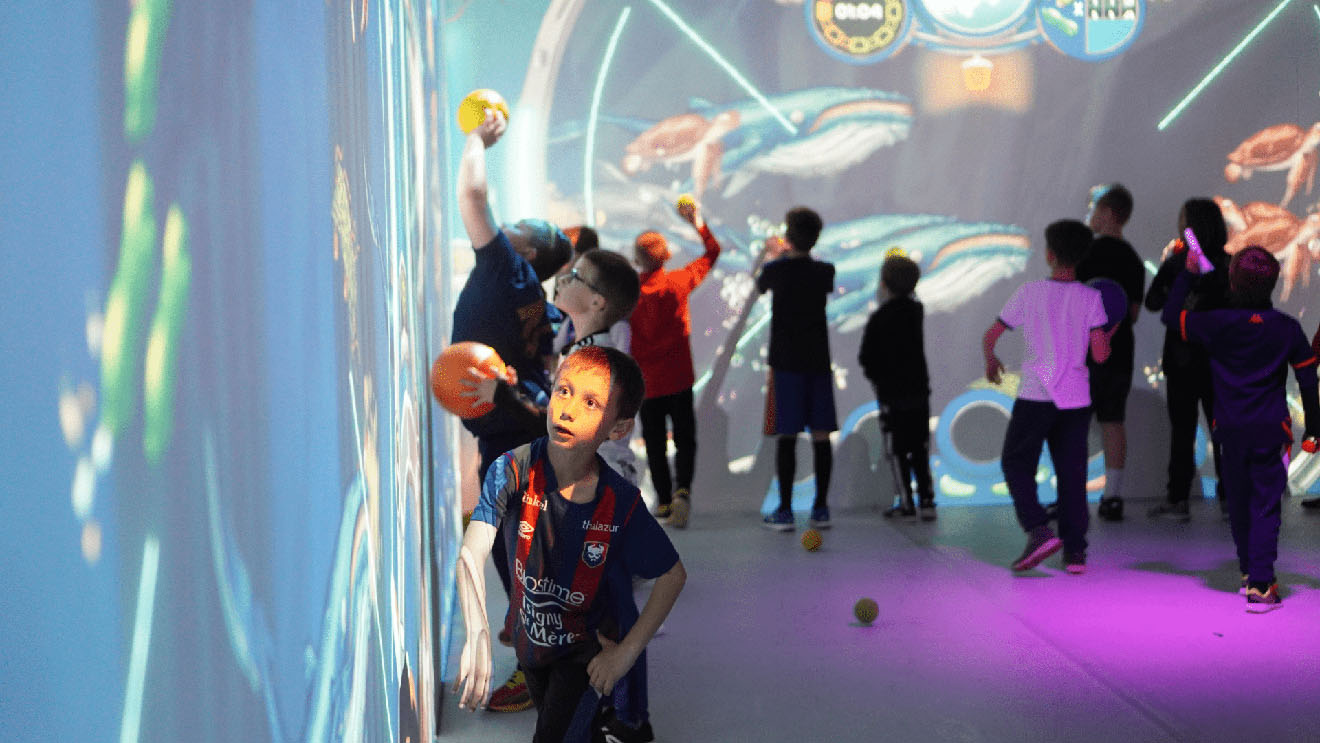 Enfants lançant des balles sur des murs interactifs affichant un jeu aquatique avec des baleines et tortues.