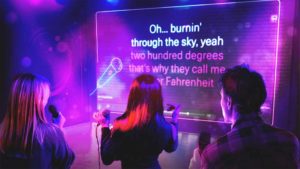Three adults singing karaoke with microphones, in front of a screen displaying the lyrics