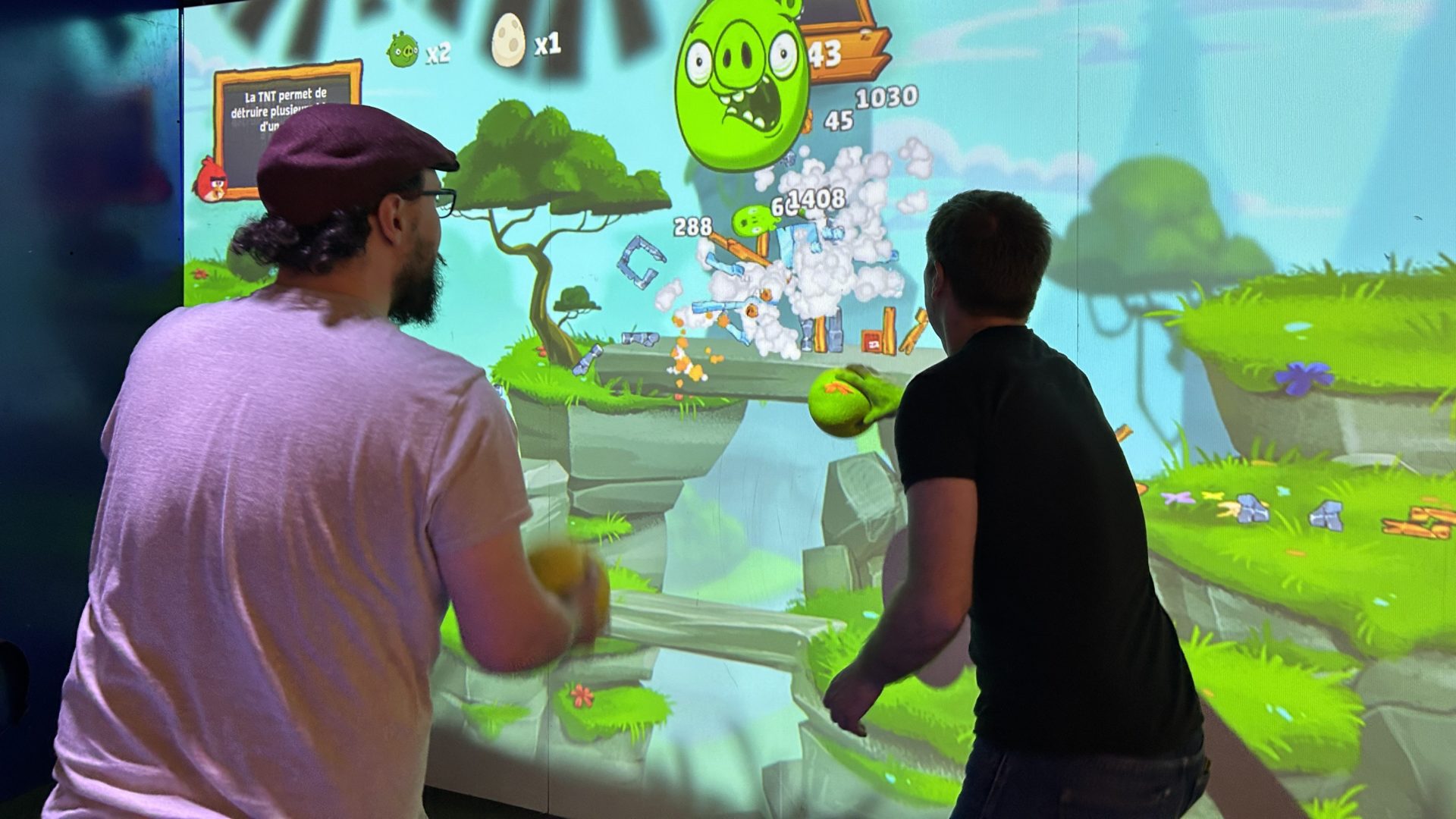 Deux adultes lancent des balles sur un mur interactif en jouant à Angry Birds Smash, avec une explosion de points et d’éléments à l’écran